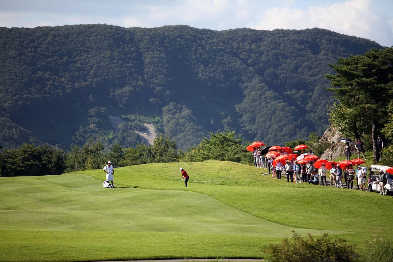 A golf tournament in progress: a player chipping onto the green while spectators with umbrellas watch from the bank